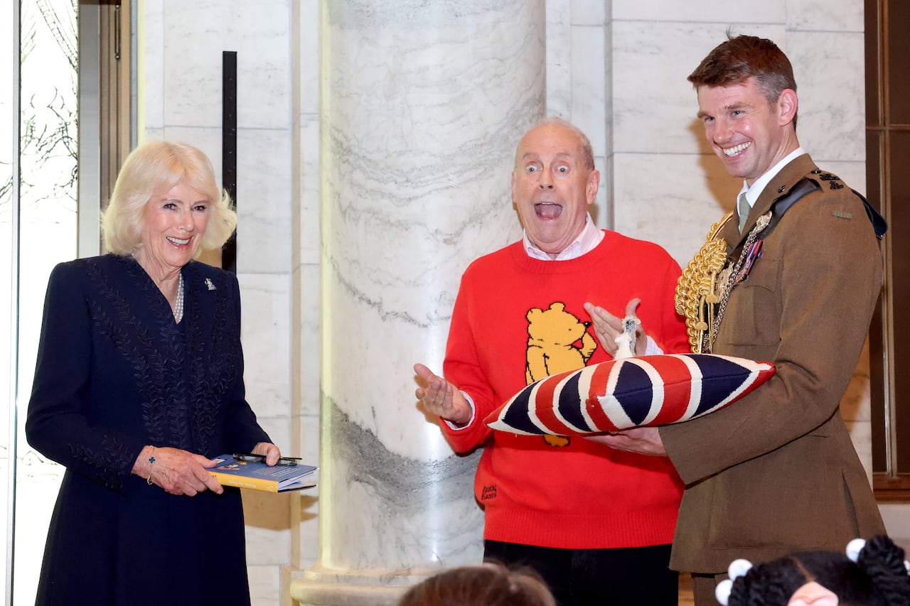 A blond woman in a dark blue dress holds a book and smiles as a bald man in a red sweater with Winnie-the-Pooh on it opens his mouth wide in an expression of surprise as a man in uniform holds a union jack pillow with a small stuffed toy on it.