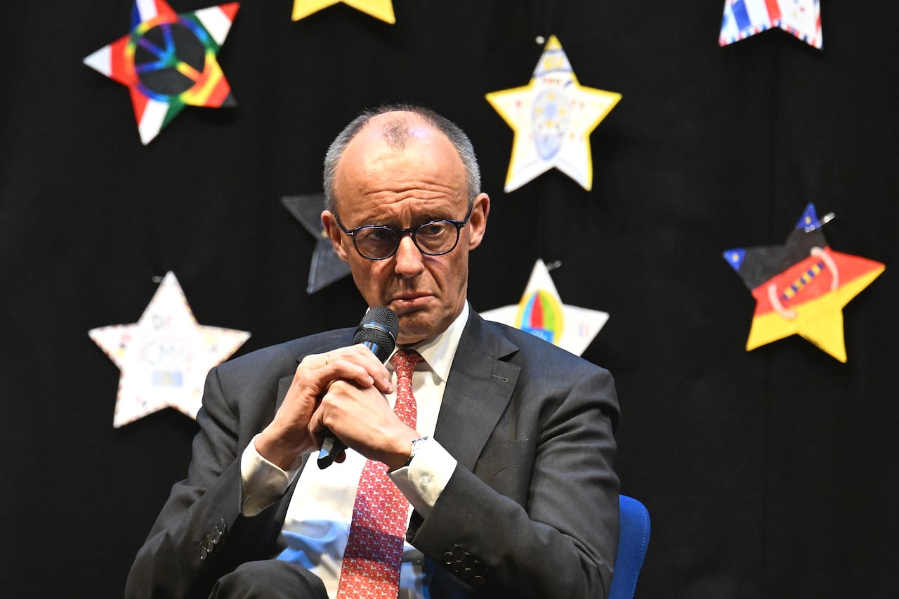 A bespectacled, balding man in a suit and tie holds a microphone in two hands while speaking.