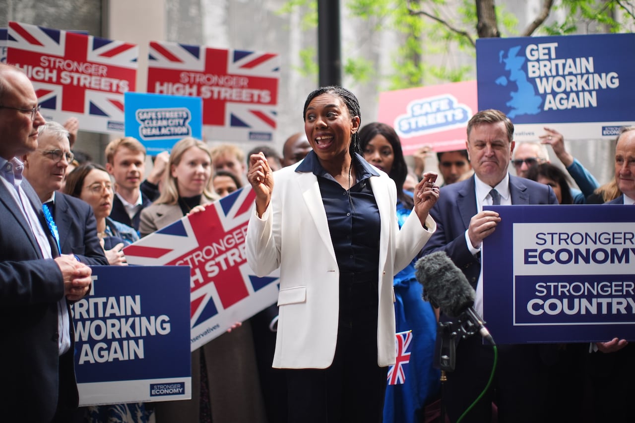 A woman in a white blazer smiles while talking to reporters and a crowd holding signs that say, 'Get Britain working again.'