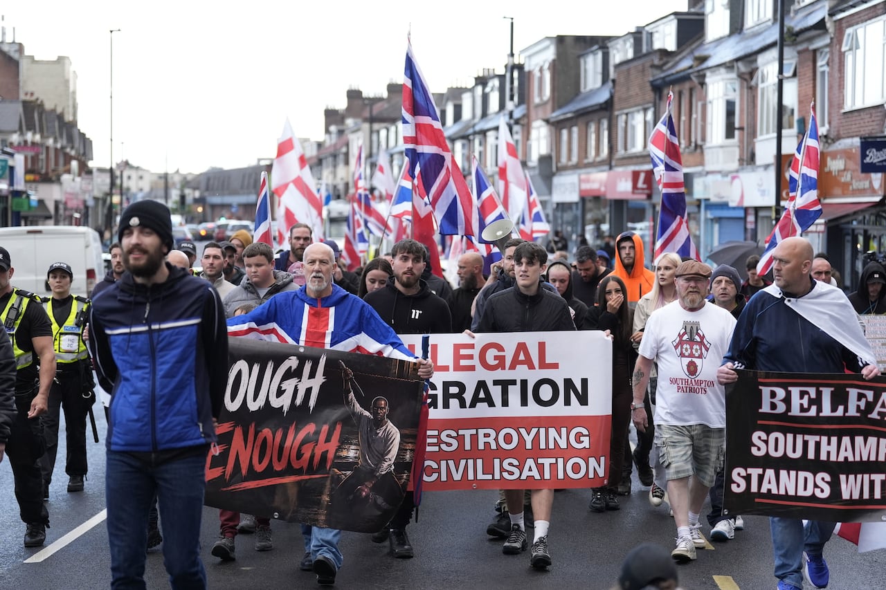 Dozens of people are shown walking down the street holding signs and flags in an outdoor demonstration.