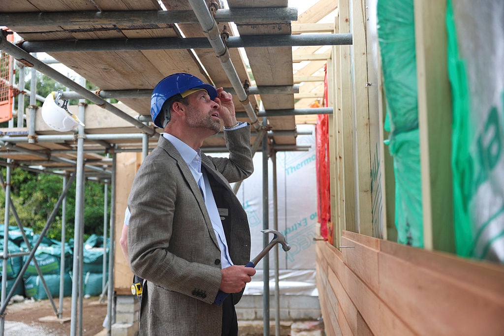 A person holds their left hand to their hard hat and a hammer in their right hand as they look up at a wall in a home under construction.