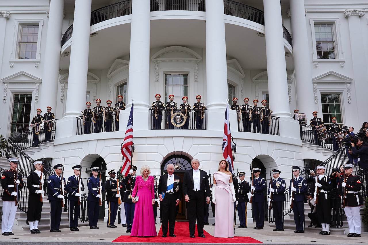 Four people in formal eveningwear stand on a red carpet in front of the White House, flanked by uniformed soldiers standing at attention.