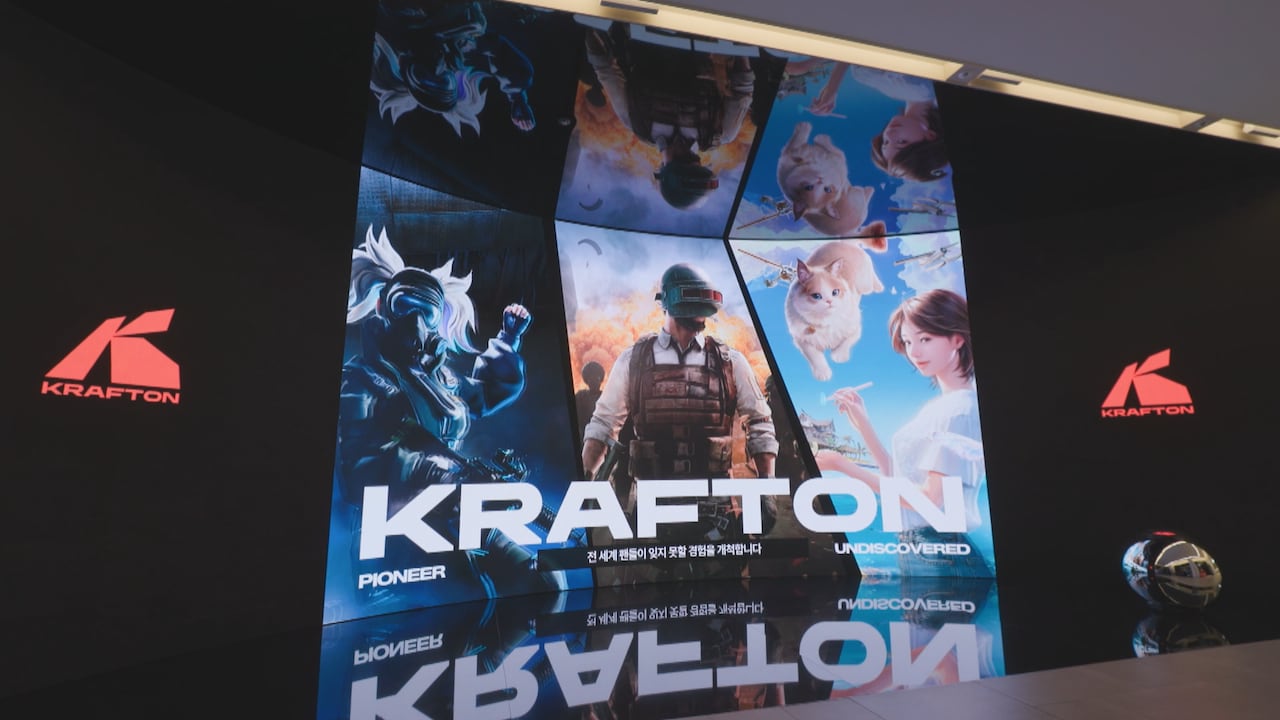 A concave screen shows the logo of Krafton, a videogame company, along with three images of their games -- a white haired character in a mask, a soldier-like character in a mask, and a woman in white with a cat floating on top.