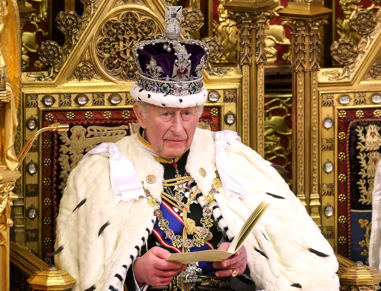A person wearing a crown sits on an elaborately appointed seat holding a speech they are reading.