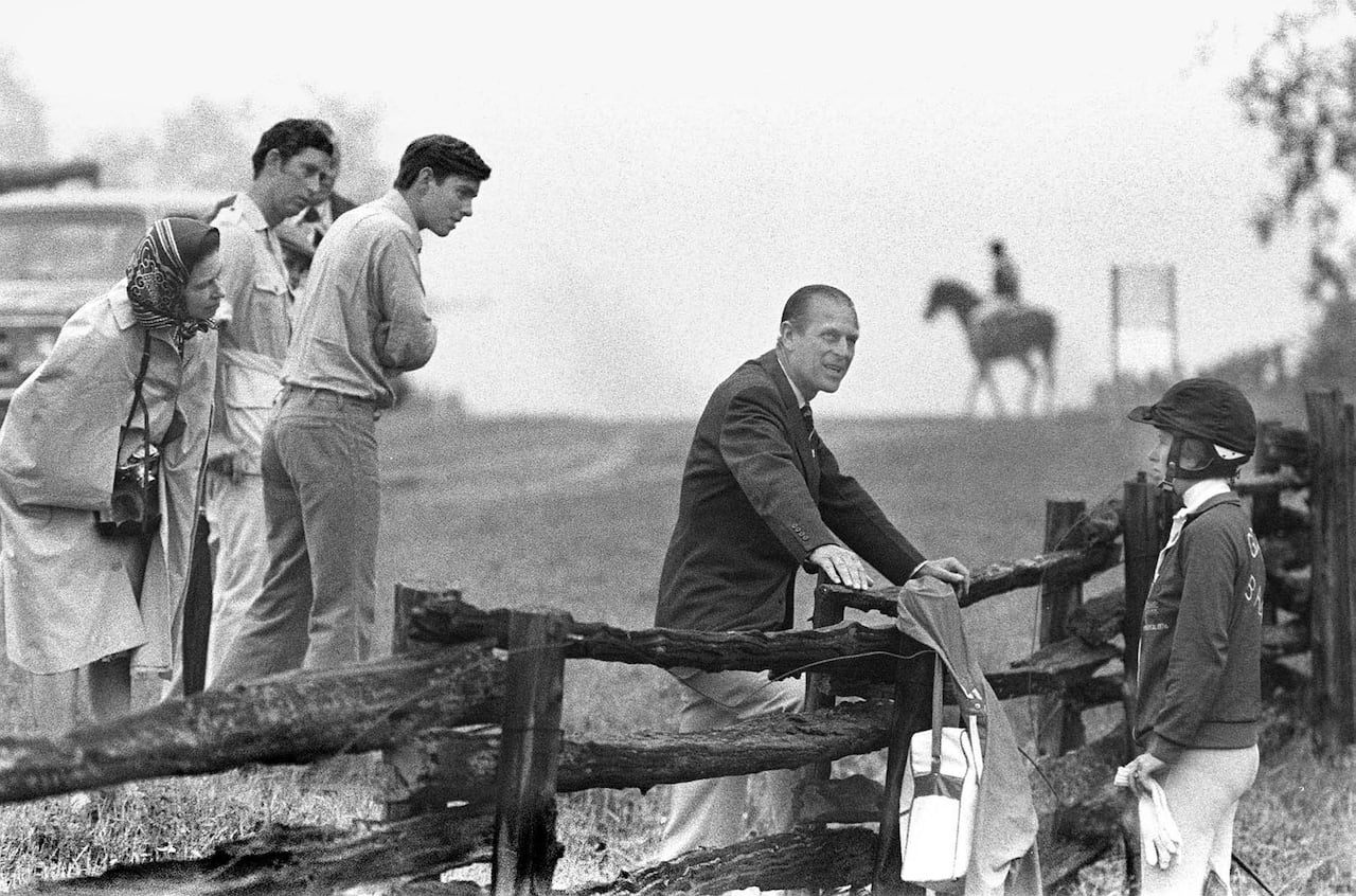 A person leaning on a fence talks to another person on the other side as three people watch and a horse and rider are in the background.
