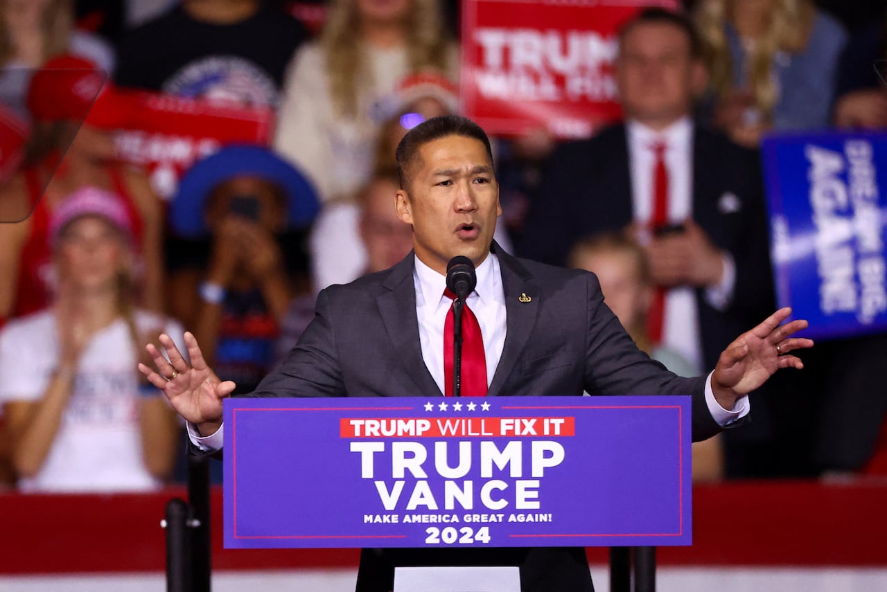 Man in dark suit speaks to a crowd from a podium with a banner reading "Trump will fix it."