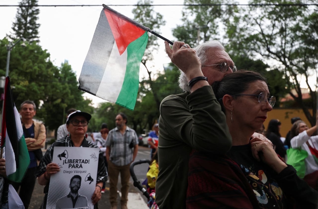 People are shown at an outdoor demonstration, men and women, holding signs and a multicolored flag.