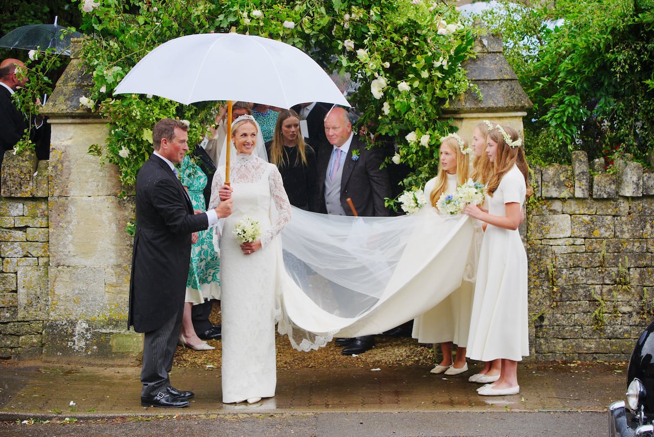 A bride and groom stand on a sidewalk and three bridesmaids hold the bride's train.