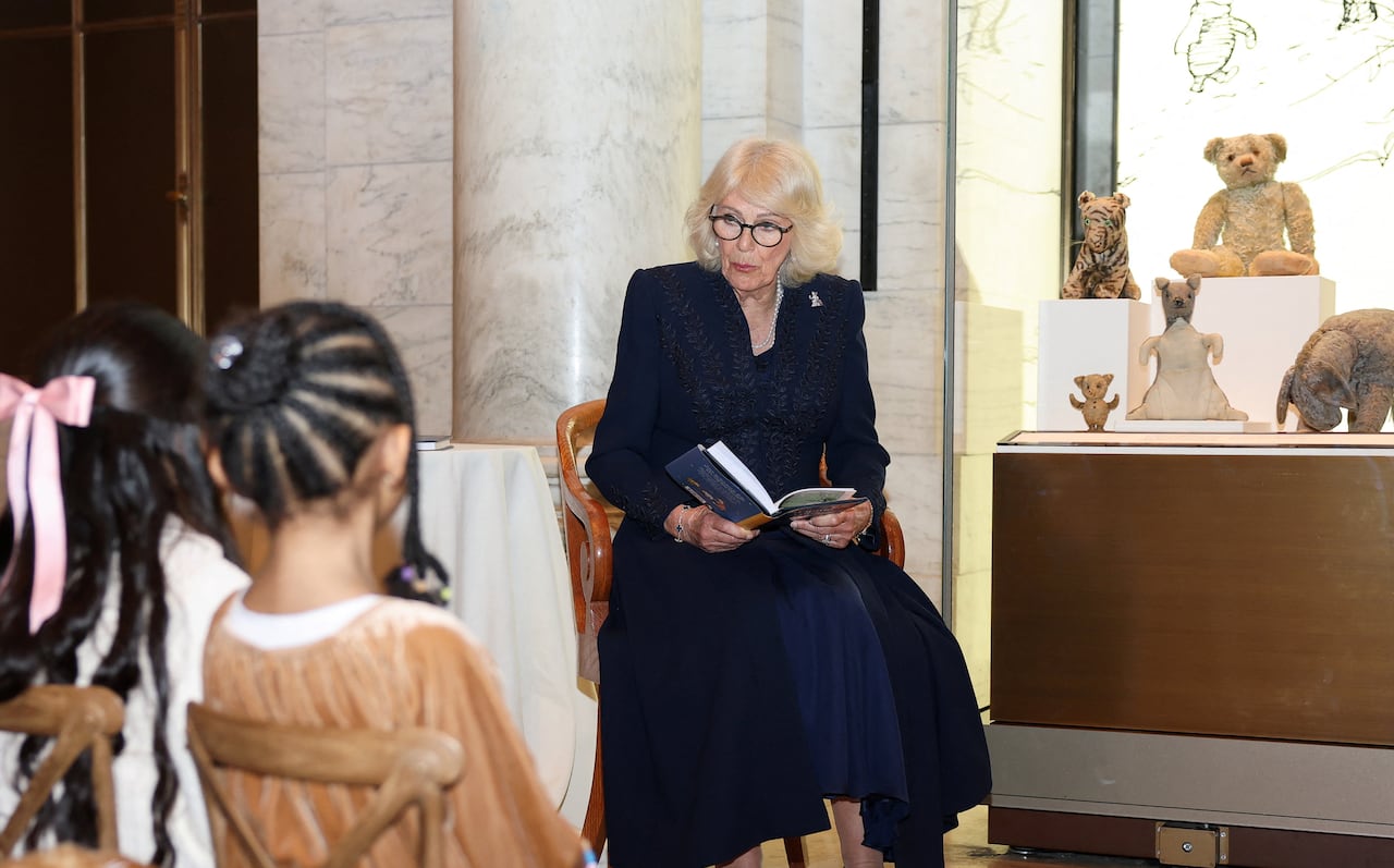 A blond woman in a blue dress sits on a chair reading a book to several children seated in front of her. She is flanked by a museum case filled with stuffed animals.