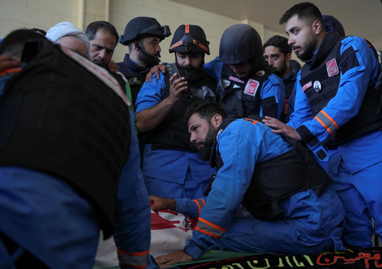 A group of men wearing blue paramedic uniforms, some wearing helmets, embrace around the shroud-covered body of a man on the ground.