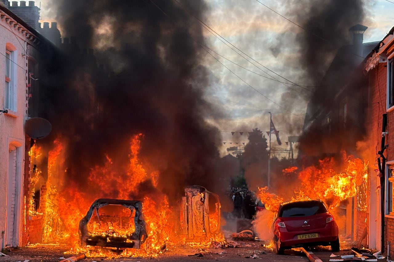 Huge orange flames are shown burning at least two vehicles on a narrow street as well as low-rise buildings.
