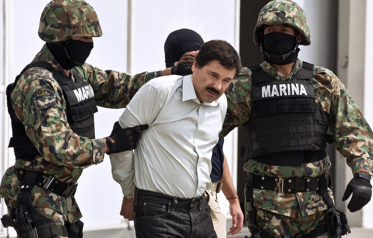 A man with a mustache and a button up dress shirt stands between two soldiers, with one, on the right, pushing his head down slightly.