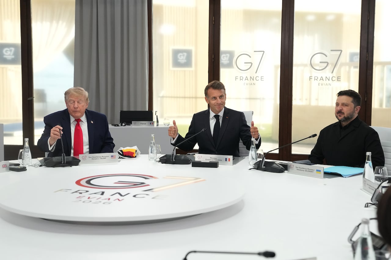Three men are shown seated at a large circular table. Two are cleanshaven and wearing suit and tie, the other is bearded and wearing dark clothing.
