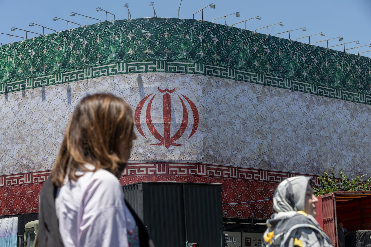Women are seen walking past a billboard displaying the Iranian flag.