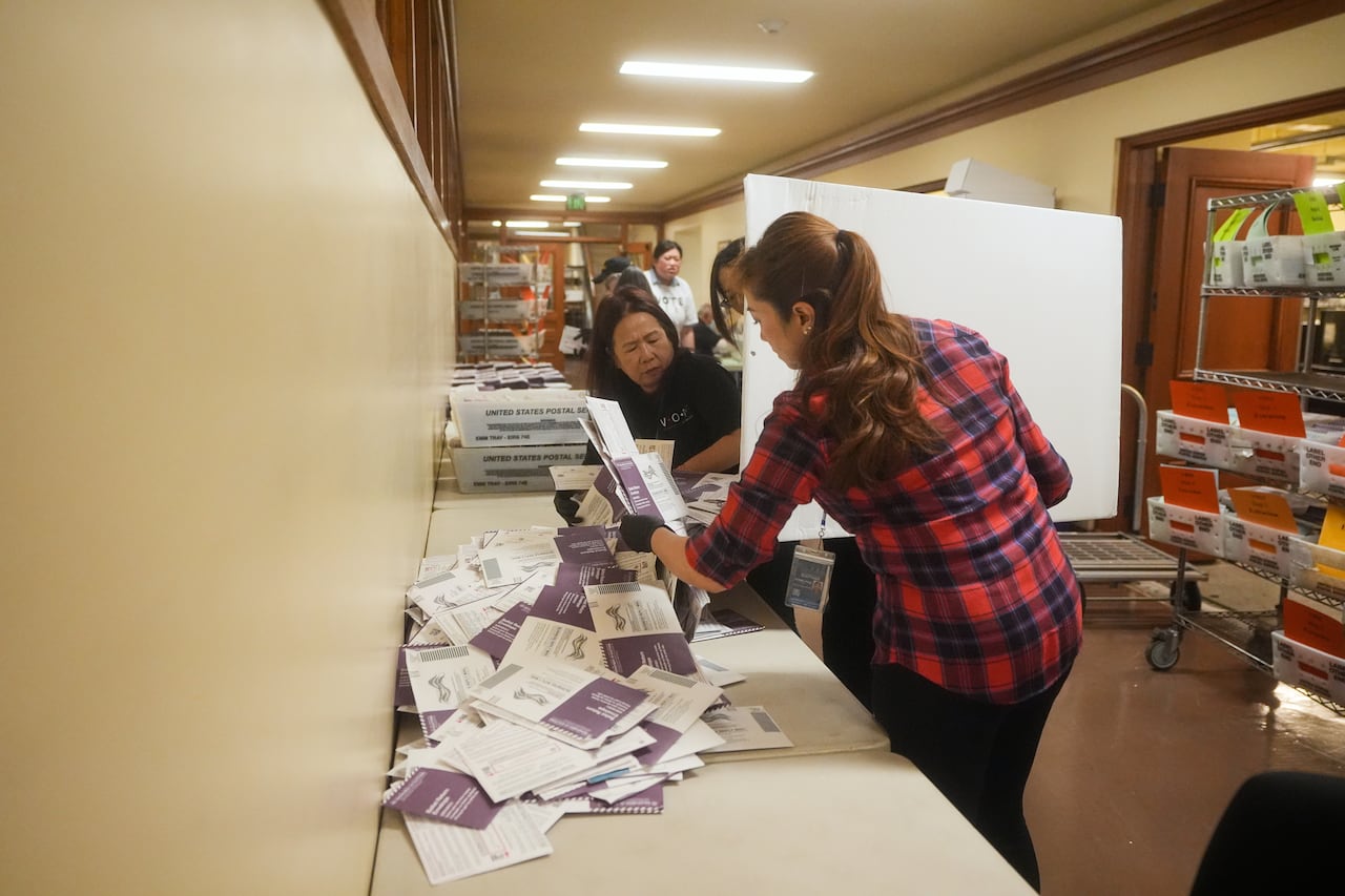 Two women are shown in the foreground near several pieces of paper, which appear to be ballots.