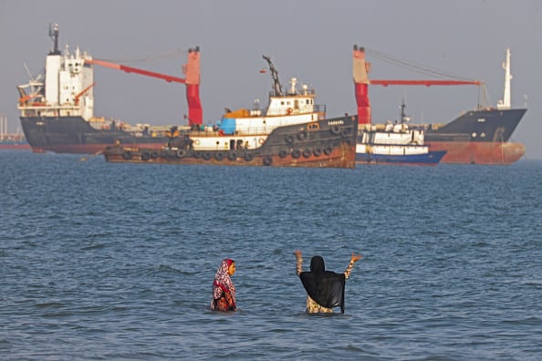 Two women, clothed, swim at a beach along the Strait of Hormuz. Cargo ships can be seen in the distance in the sea.