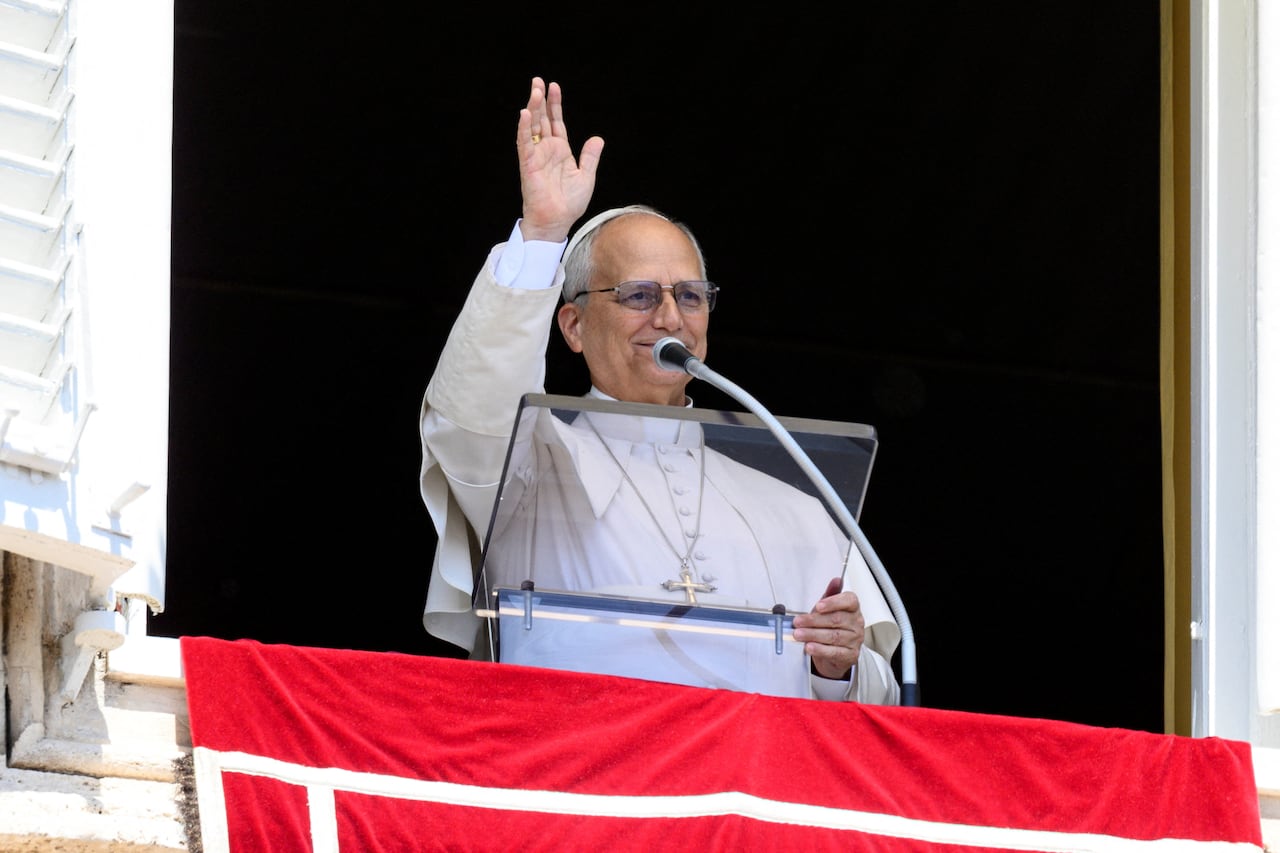 An older man in white robes speaks from a balcony