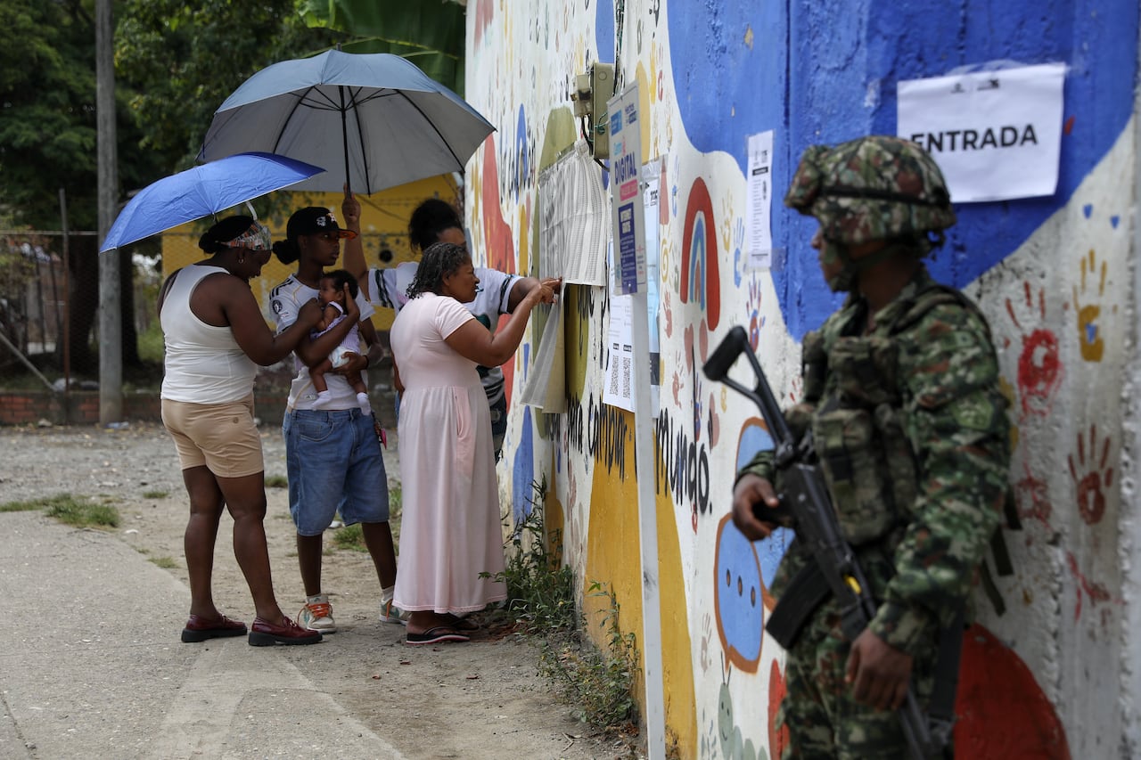 A soldier stands guard as people look at papers posted on a wall.