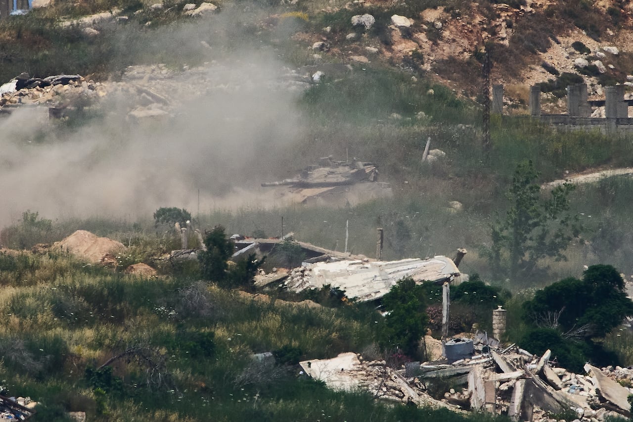 A tank kicking up dust amid damaged structures is seen from afar.