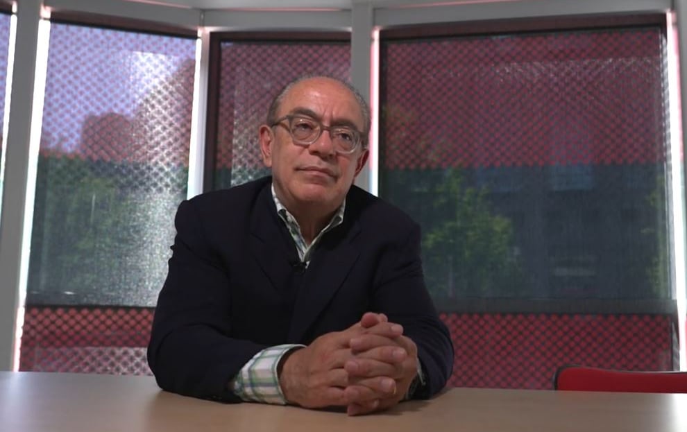 A man with glasses sits a desk with hands clasped.