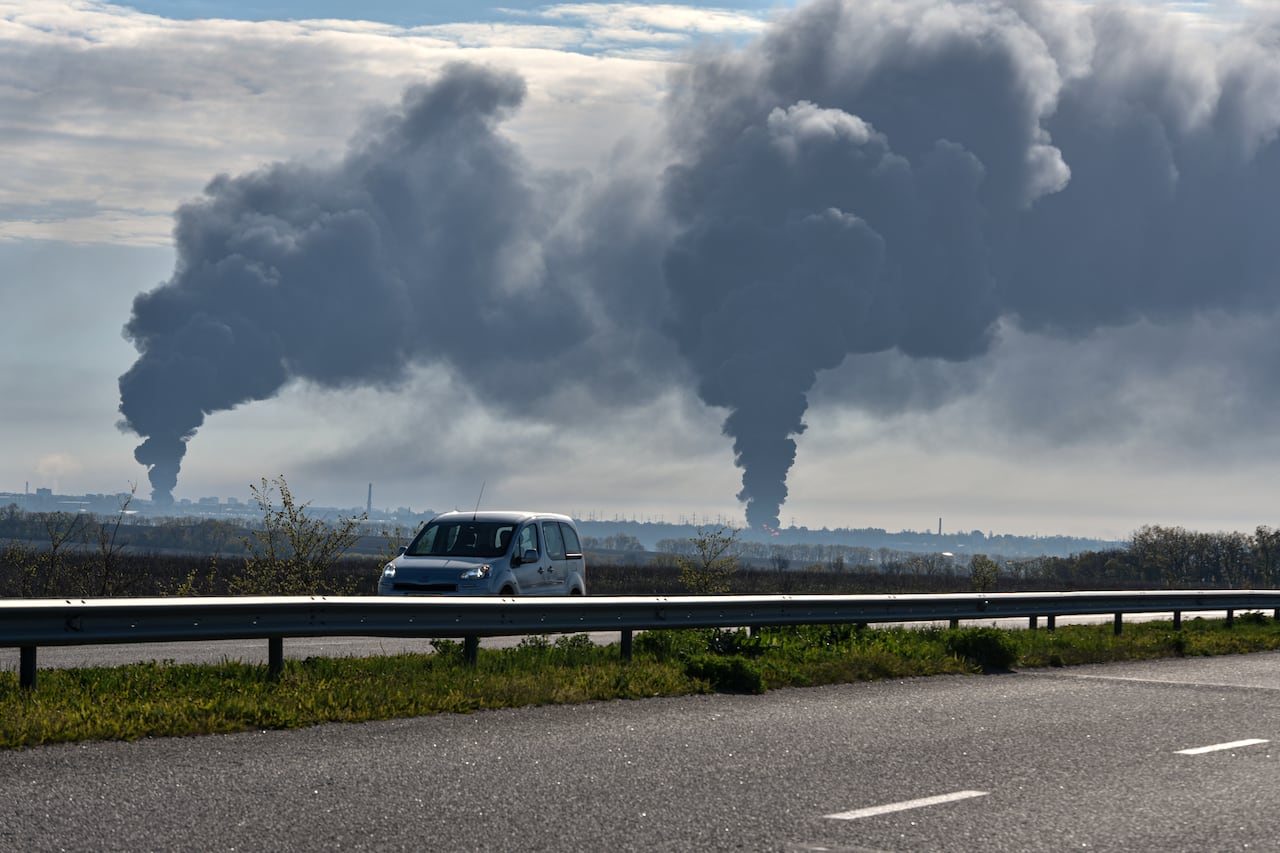 A car drives on a highway with billows of smoke rising in the distance behind it