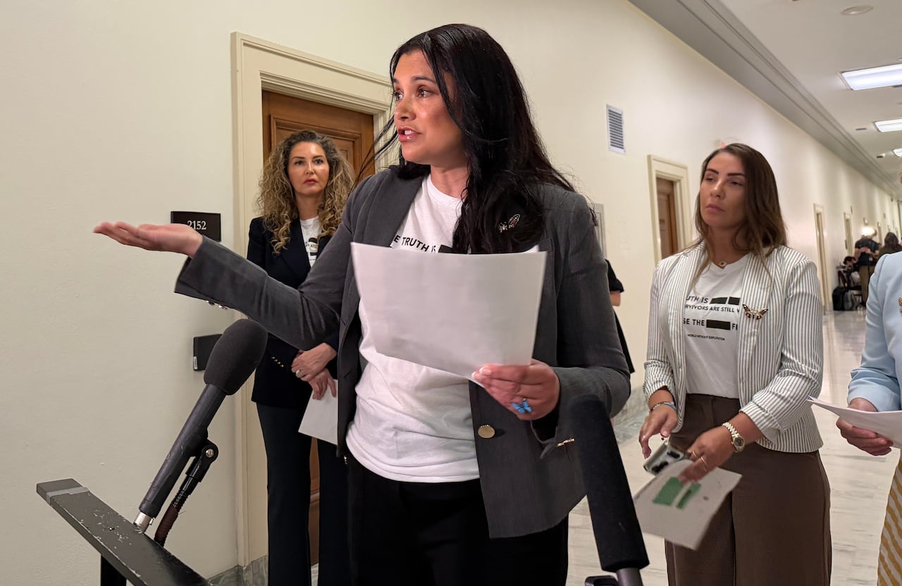A woman holding a sheaf of papers speaks at a microphone, while standing in a hallway. Other women look on.