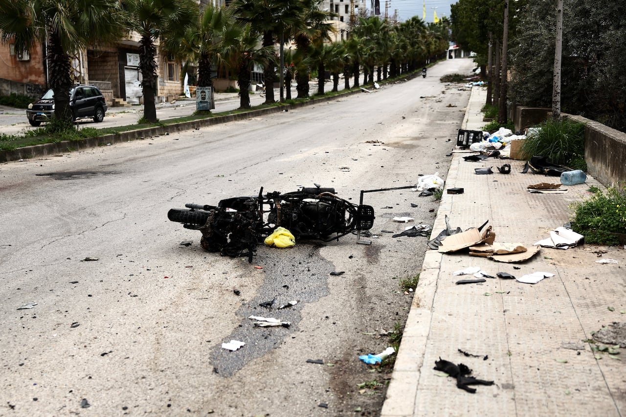 The charred remains of a motorized scooter lies in a palm tree-lined street among pieces of trash.