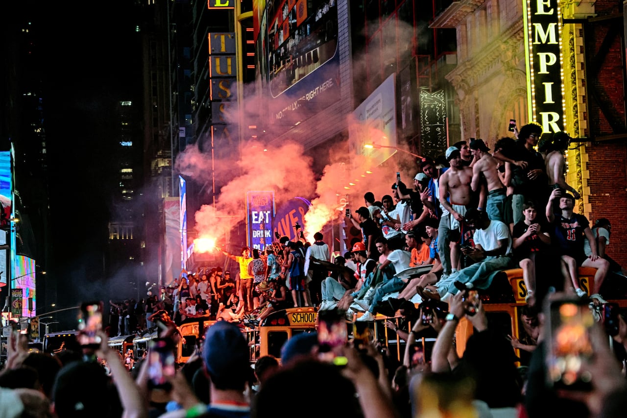 Orange flares rise above crowds filling a street lit by many signs and people standing on top of a school bus.