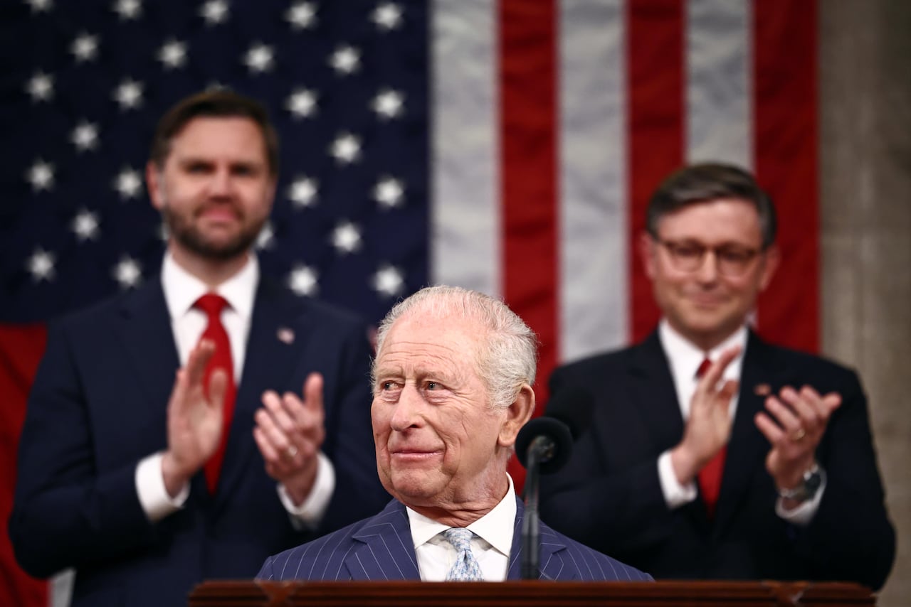 An older man with silver hair wearing a navy pin-striped suit stands at a podium as two men in dark suits and red ties stand in front of an American flag and applaud.