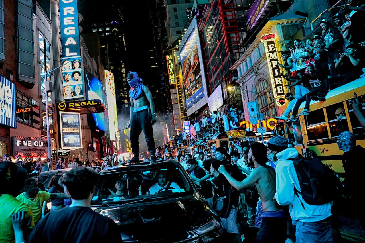 Fans fill a large city street brightly lit by signs. One person stands on top of a car with their shirt lifted above their navel.