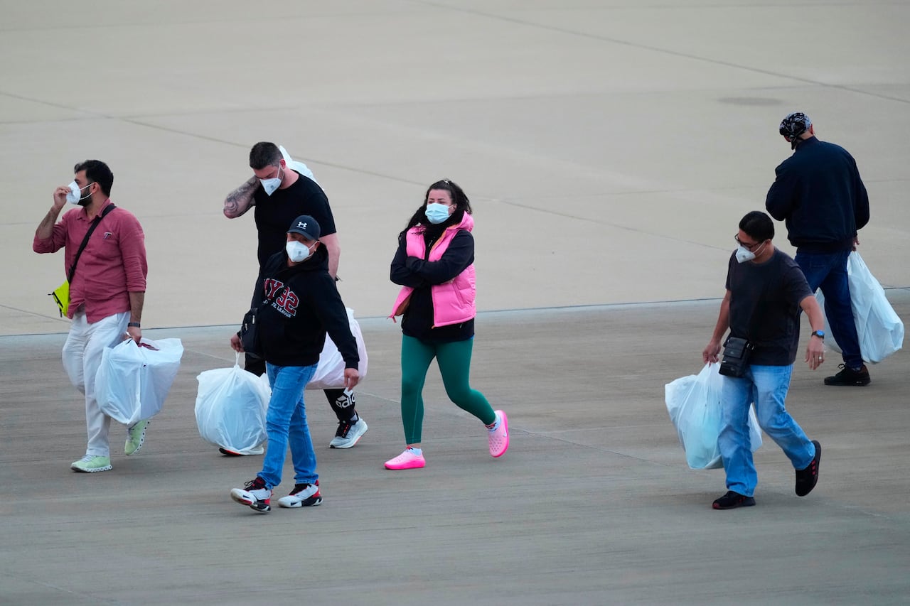 Several people, men and women wearing masks over their nose and mouth, are shown walking on a tarmac.