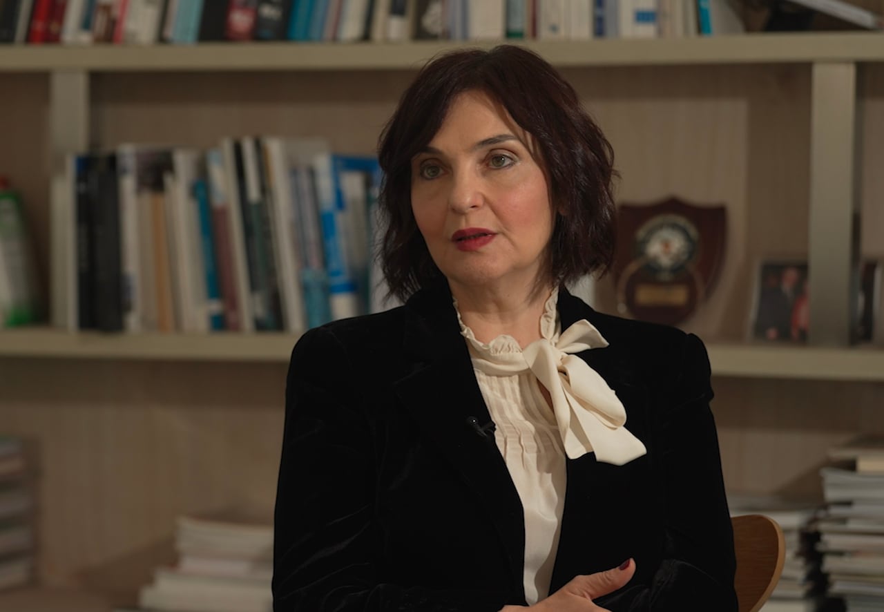 A woman with shoulder length dark hair wears a dark jacket and a white blouse as she speaks while sitting in front of a book case.