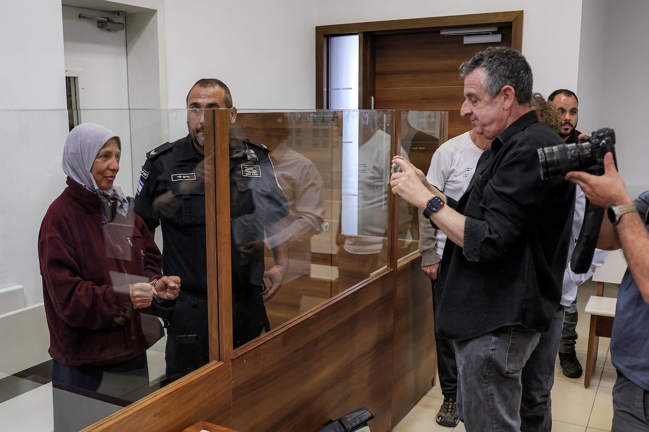 A women in a head covering is shown behind a glass partition in what appears to be a courtroom, with photographers on the other side taking her picture.
