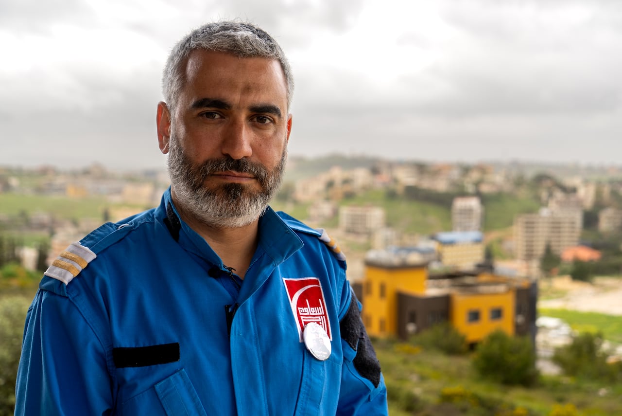 A man with greying hair and a beard wearing a blue paramedic's uniform looks at the camera. Buildings and greenery can be seen out of focus behind him.