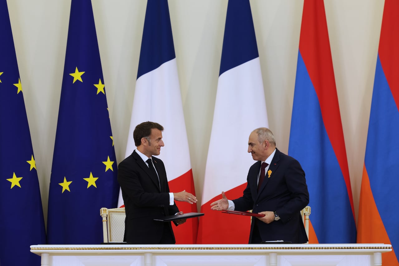 Two white men reach in to shake each other's hands at a podium in front of many large flags.