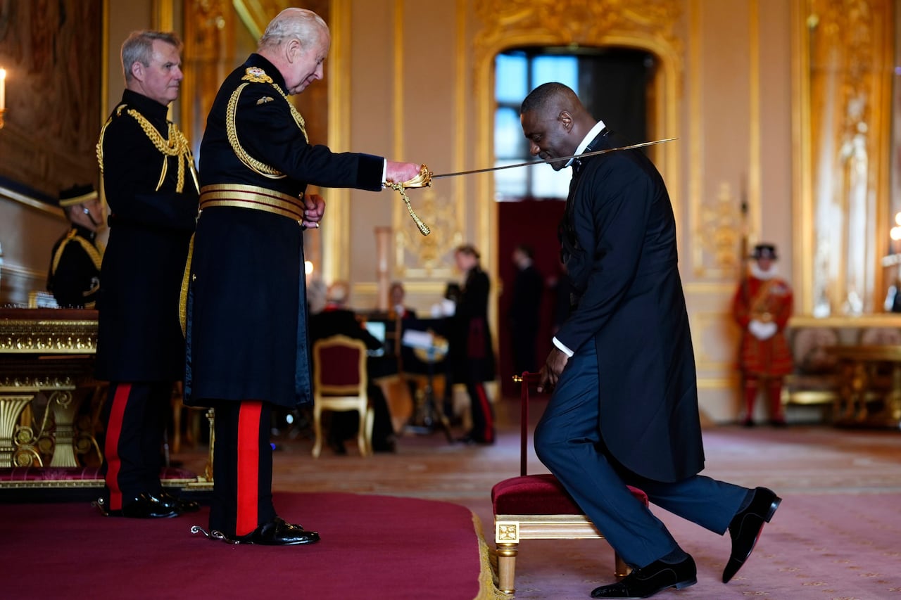 A person taps a sword on the shoulder of a person kneeling in front of them.