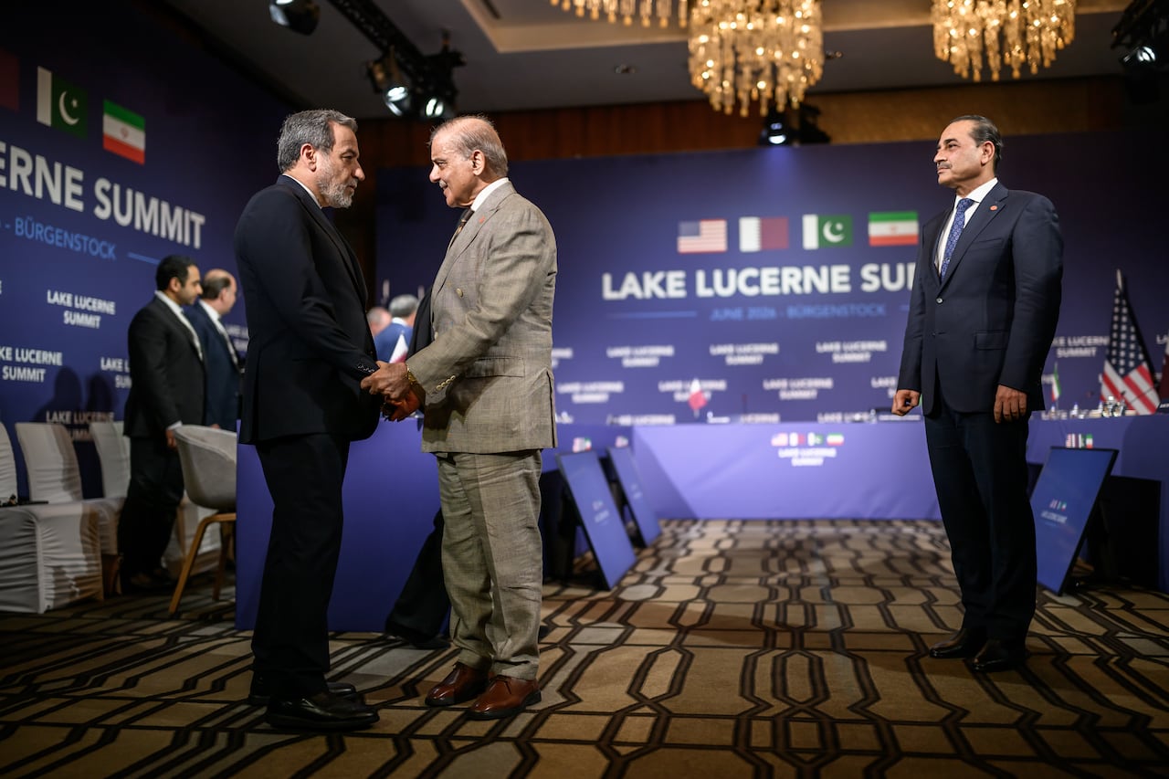 Two men in suits shake hands while another man looks on.