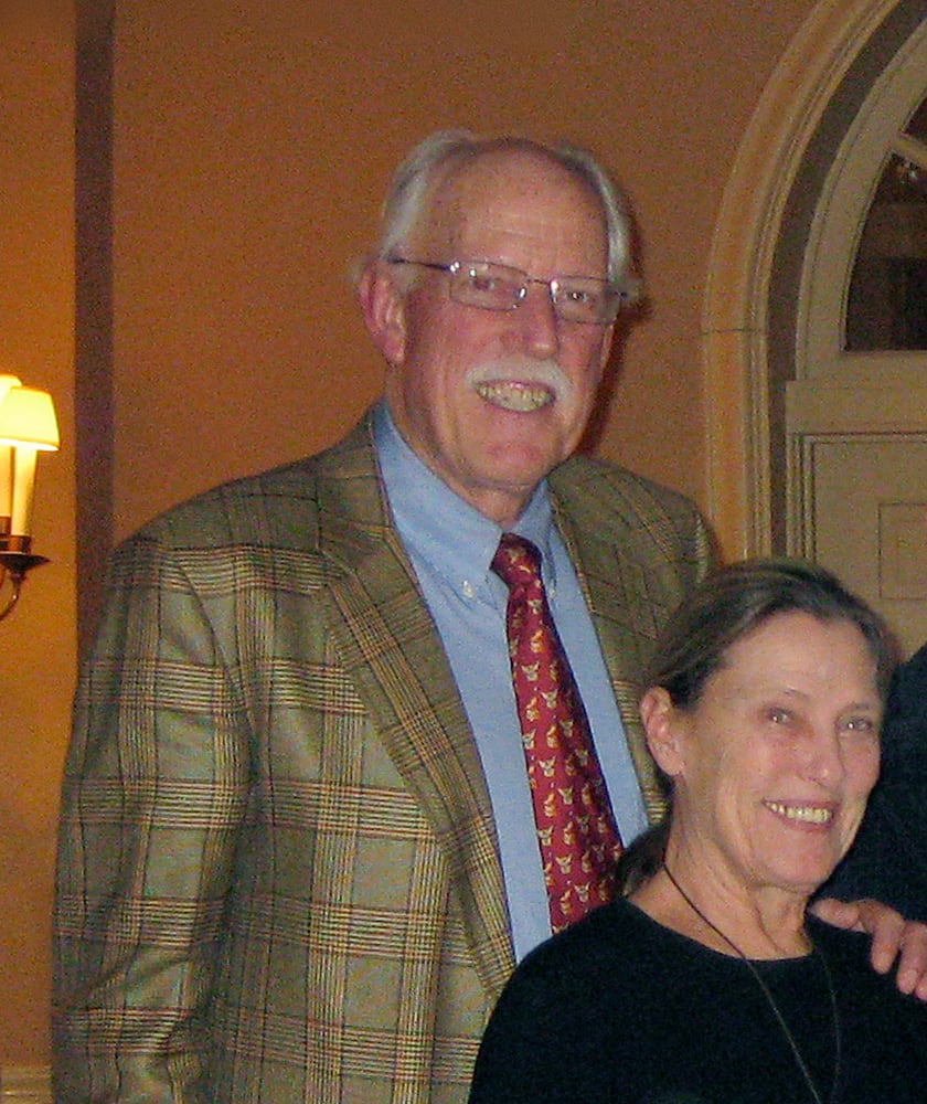 An older man with white hair and a white moustache wearing a brown plaid jacket smiles as he stands behind a smiling woman wearing a black sweater with her long, brown hair in a ponytail.
