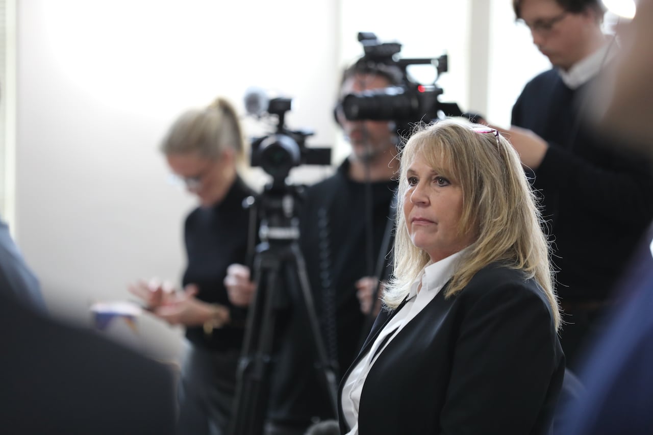 An older woman with blonde hair and in a dark suit looks to the left, with memebers of the media and videographers seen behind her.