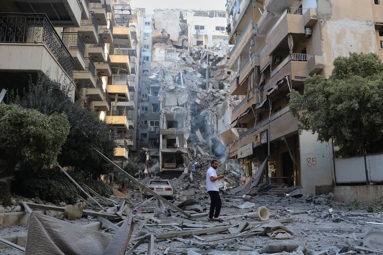 A man is shown from a distance standing on a street filled with concrete and other debris as multistorey buildings are shown damaged all around him.