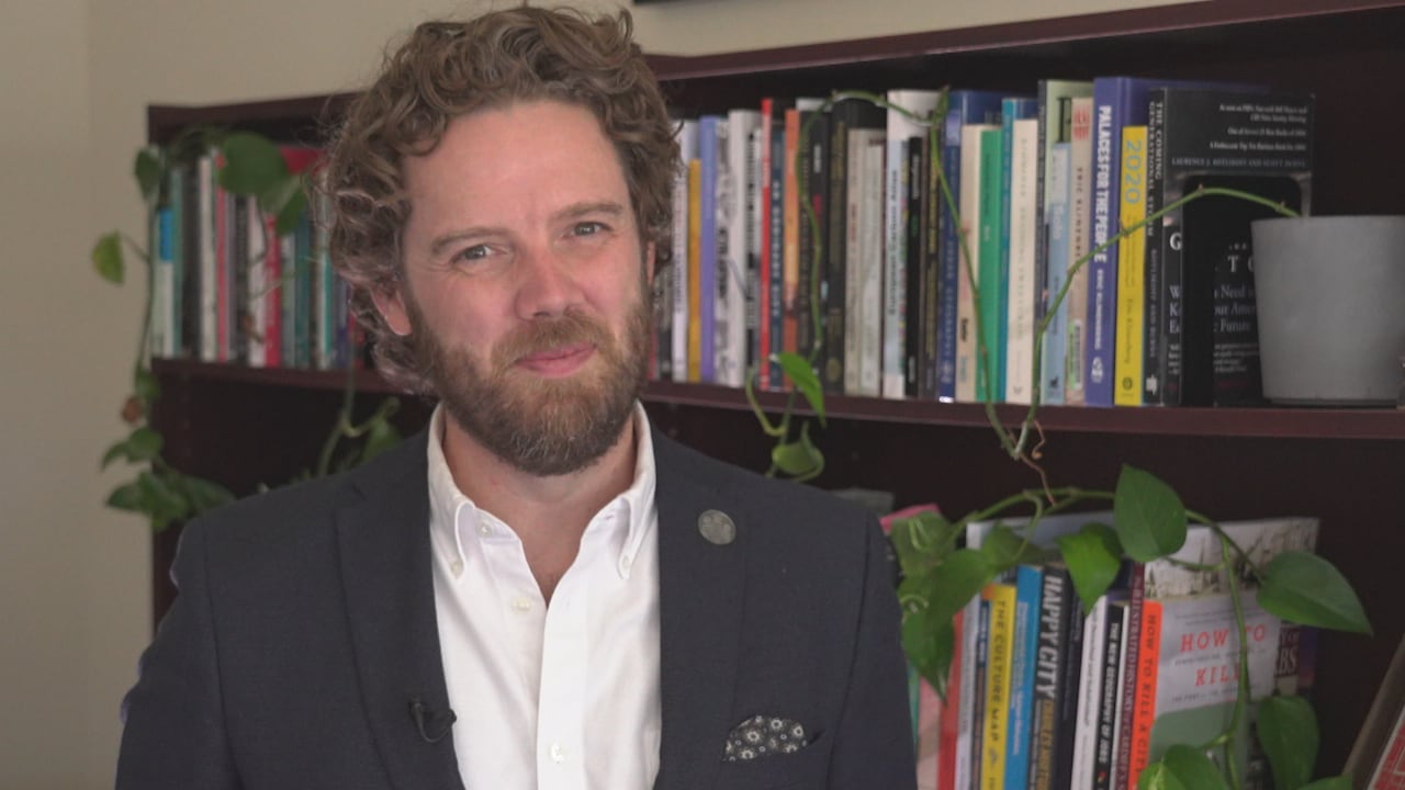 A man in a suit smiles at the camera with a bookcase and plants behind him.