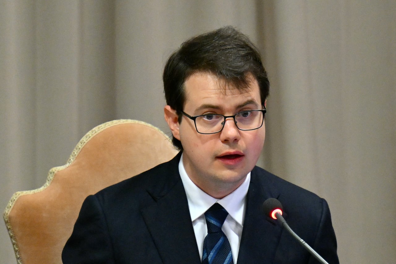 A bespectacled, cleanshaven younger man in a dark suit and tie speaks and is shown in closeup.