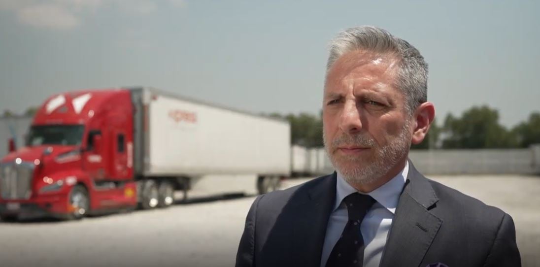 A man with a suit stands with a transport truck in the background.