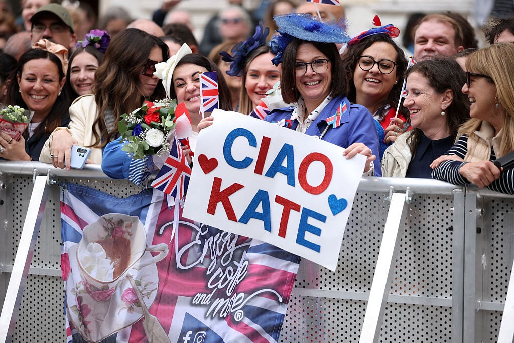 People stand behind a barricade, with one holding a sign that says 'Ciao Kate.'