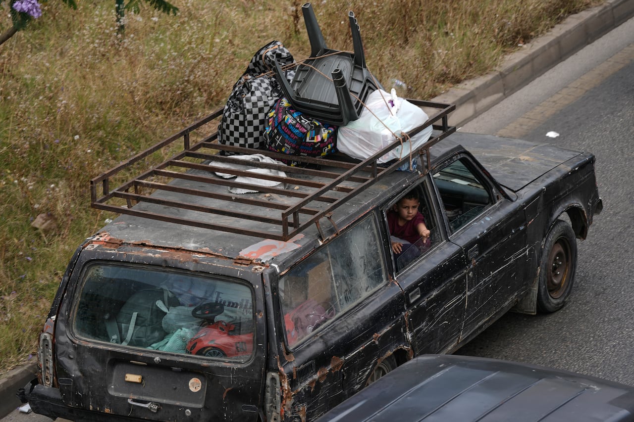From an aerial view, a rusty and old looking vehicle is shown with items strapped to its roof. A boy can be seen in the car through an open window.