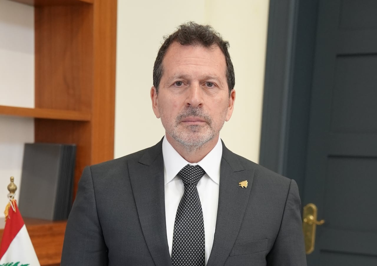 A man in a suit and tie stands in an office flanked by a Lebanese flag.