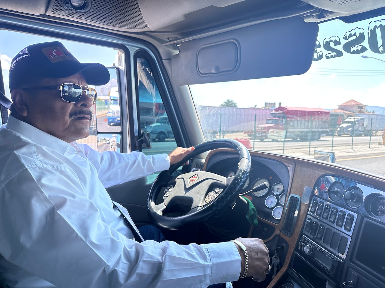 A man with a baseball cap and sunglasses sits in the cab of a transport truck.