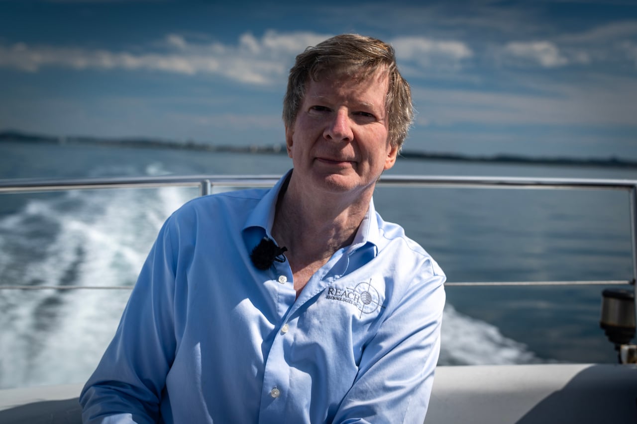 Glenn Jones is a white man in his 60's wearing a light blue button up shirt that says Reach Technologies on the chest. He's sitting on a boat in the middle of the pacific ocean.