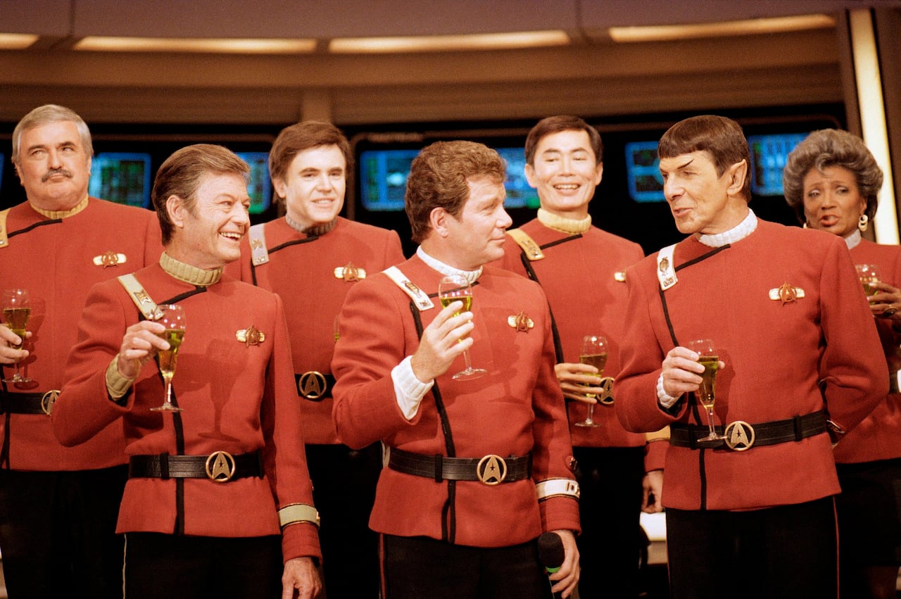 Seven actors in red sci-fi style uniforms post for a photo, holding glasses of wine.