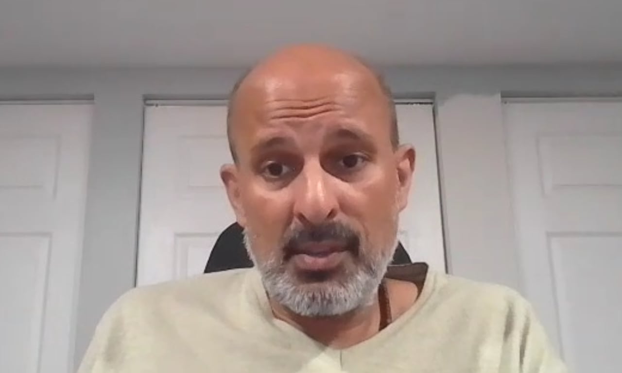 A Zoom screenshot of a man sitting in a white and grey room